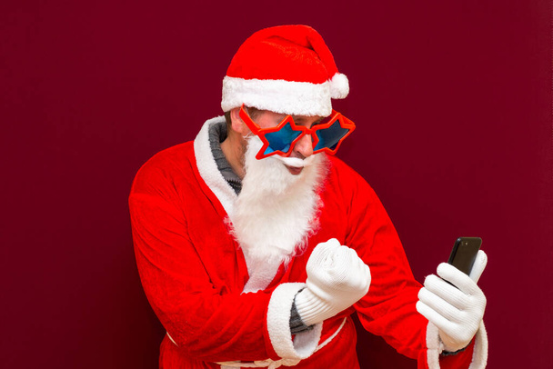 Πορτρέτο του trendy Santa Claus χρησιμοποιούν το κινητό τηλέφωνο και σφιγμένα fistswearing μοντέρνα αστέρια γυαλιά γυαλιά καπέλο ανάγνωση ειδήσεις απομονώνονται πάνω από το κόκκινο φόντο - Φωτογραφία, εικόνα