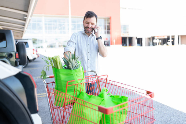Lächelnder hispanischer Mann spricht mit Smartphone, während er mit Einkaufswagen im Auto steht - Foto, Bild