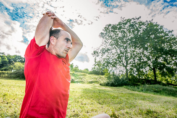 Homme athlétique faisant étirement dans la nature avec masque et chemise rouge - Photo, image