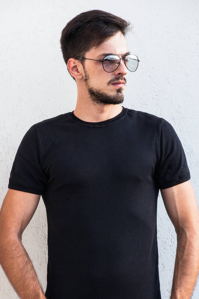 młody człowiek w okularach w czarnej koszuli outdoor shot w przedniej białej ścianie t shirt mock up - Zdjęcie, obraz