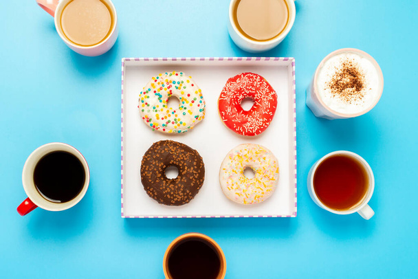 Tasty donuts and cups with hot drinks, coffee, cappuccino, tea on a blue background. Concept of sweets, bakery, pastries, coffee shop, meeting, friends, friendly team. Flat lay, top view. - Photo, Image