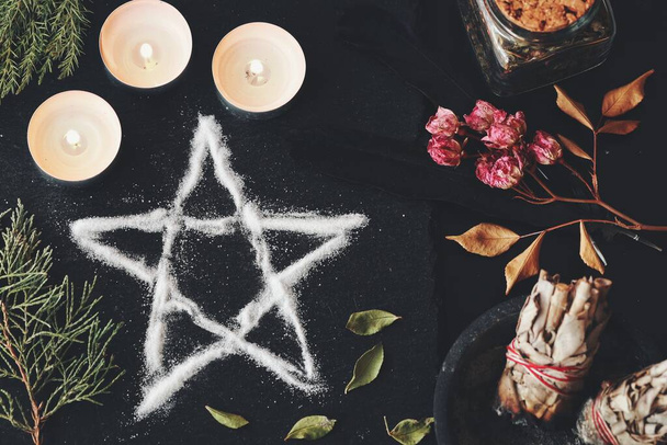 Salt pentagram symbol on wiccan witch altar. Flat lay of hand made pentagram for protection and meditation for witchcraft ritual. Candles, dried flowers, sage smudge sticks evergreens on black surface - Photo, Image