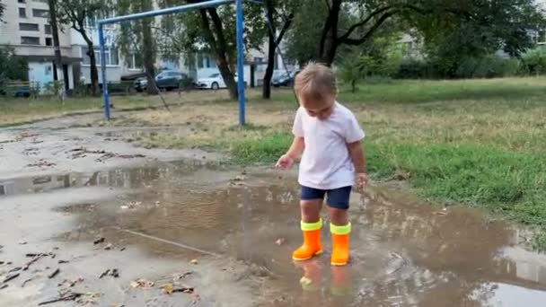Kleinkind in orangefarbenen Stiefeln geht nach dem Regen durch die Pfützen. - Filmmaterial, Video