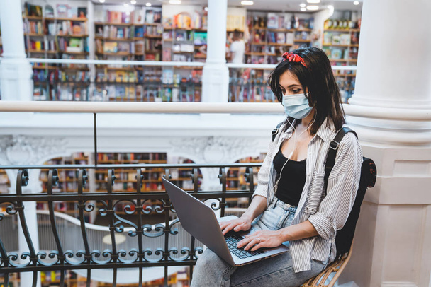 Vrouw zit op een stoel en werkt aan een laptop in de bibliotheek. Meisje met beschermend gezichtsmasker studeert aan de universiteit tijdens wereldwijde pandemie - Foto, afbeelding