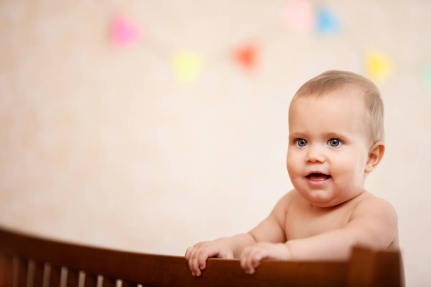 Sellective focus of happy little baby standing in wooden crib indoors, decorated nursery. Side view of lovely naked caucasian child laughing with mouth open. Concept of happychildhood. - Foto, afbeelding