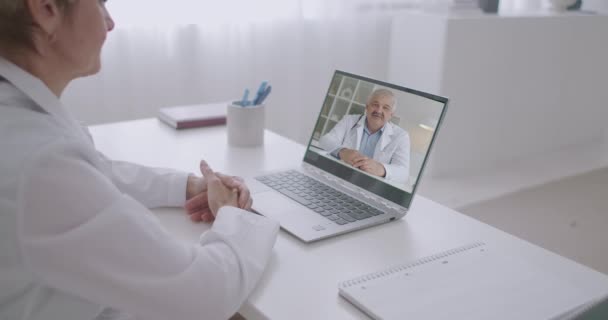 τηλεδιάσκεψη δύο ιατρών, ανδρών και γυναικών συνομιλούν μέσω web camera του laptop στο γραφείο της σύγχρονης κλινικής - Πλάνα, βίντεο