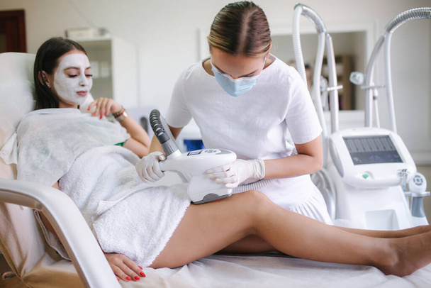 Attractive woman have a rest while cosmetologist in white gloves performing radiofrequency lifting procedure on the stomach. Beautiful woman in medical clinic. Beautician in medical mask - Photo, Image