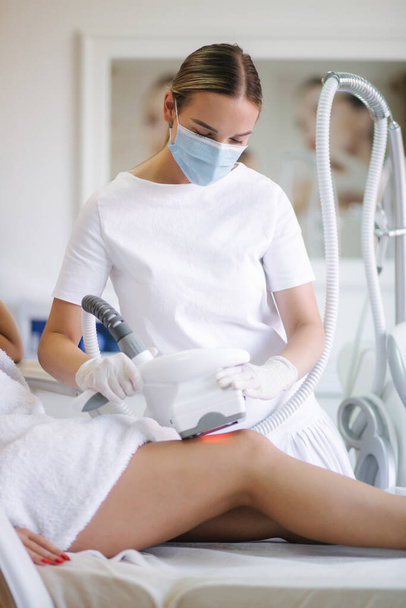 Attractive woman have a rest while cosmetologist in white gloves performing radiofrequency lifting procedure on the stomach. Beautiful woman in medical clinic. Beautician in medical mask - Fotoğraf, Görsel