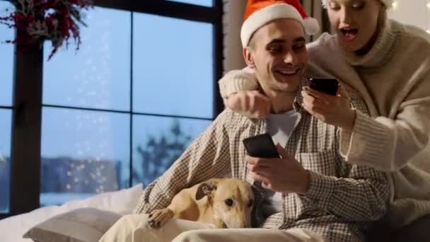 Año Nuevo. joven pareja en santa sombreros en nuevo año víspera en casa con su perro. - Imágenes, Vídeo