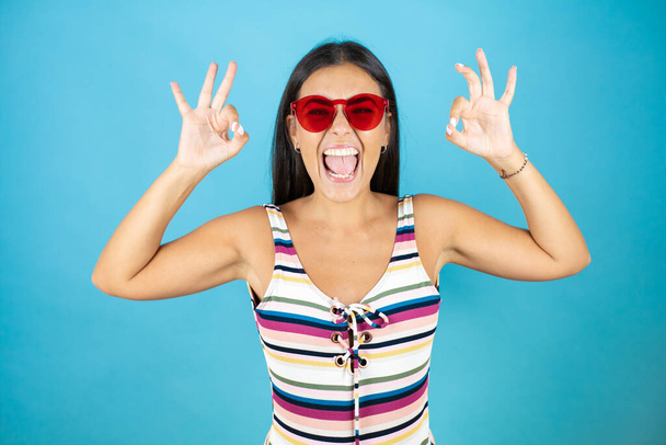 Jeune belle femme portant un maillot de bain et des lunettes de soleil sur fond bleu isolé faisant signe ok avec les doigts et souriant, excellent symbole - Photo, image