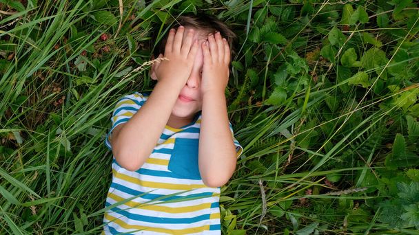 Jongen verbergt zijn ogen boven groen gras. Baby jongen bedekken sluit zijn ogen met handen en handpalmen schreeuwen lachen. - Foto, afbeelding
