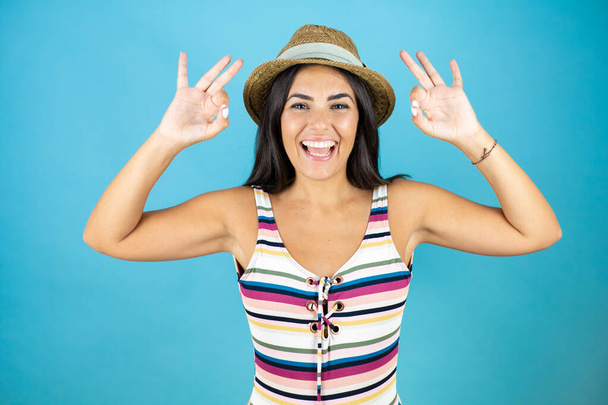 Jeune belle femme portant maillot de bain et chapeau sur fond bleu isolé faisant signe ok avec les doigts et souriant, excellent symbole - Photo, image
