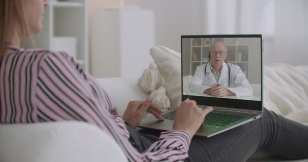 iäkäs lääkäri luennoi verkossa potilaille, nuori nainen katselee videota kannettavalla tietokoneella kotona, konsultointi ja terveydenhuolto - Materiaali, video