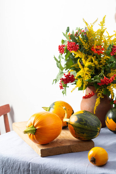 Schöner Strauß von Ragweeds, Zinnien und Viburnum in einem Tontopf, frische Ernte von Kürbissen auf einem Tisch vor einer weißen Wand - Foto, Bild