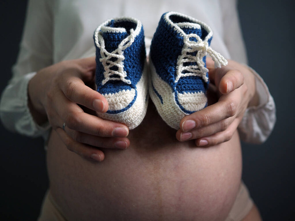 Pregnant woman holding small baby shoes relaxing at home - Photo, Image