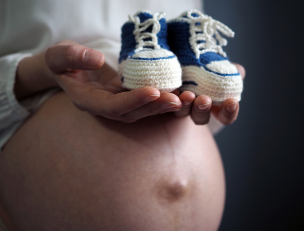Mujer embarazada sosteniendo pequeños zapatos de bebé relajándose en casa - Foto, imagen
