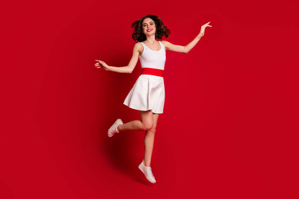 Full length photo of funny crazy lady jump high up good mood rejoicing active lifestyle lovely graceful girl wear white short dress footwear isolated red bright color background - Foto, afbeelding