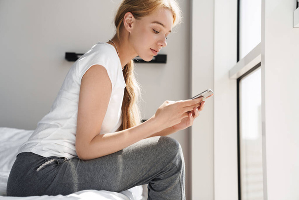 Photo of focused young woman using cellphone while sitting in bed after sleep at bright room - Photo, image