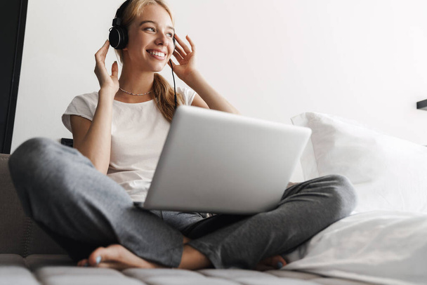 Photo of happy young woman using headphones and laptop while sitting on bed at bright room - Photo, image