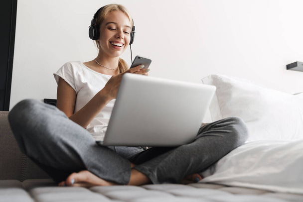 Photo of joyful young woman using headphones and cellphone while working with laptop on bed at bright room - Foto, afbeelding