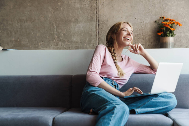 Photo of cheerful nice woman smiling and using laptop while sitting on couch in bright room - Zdjęcie, obraz