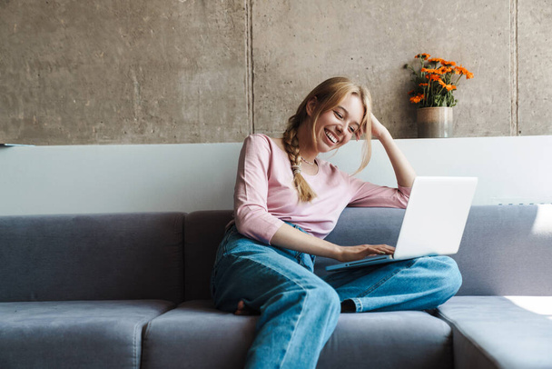 Photo of cheerful nice woman smiling and using laptop while sitting on couch in bright room - Фото, изображение