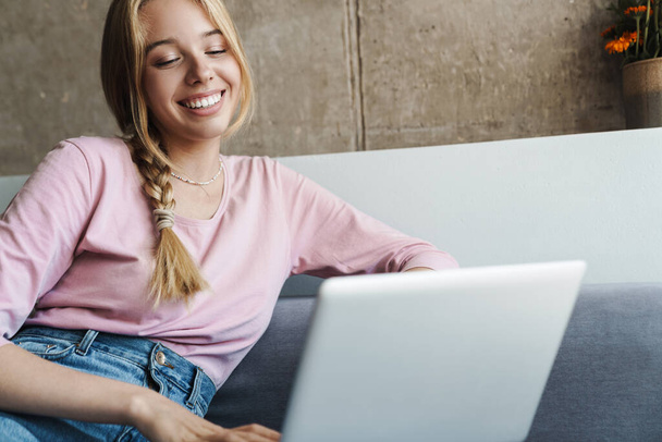 Photo of cheerful nice woman smiling and using laptop while sitting on couch in bright room - Φωτογραφία, εικόνα