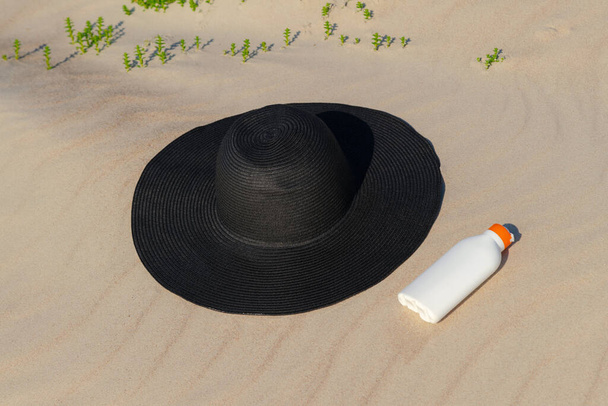 Naptej krém mellett egy fekete kalap a homokban egy napsütéses napon a strandon. Kin védelem kozmetikai napozáshoz. Senki. - Fotó, kép