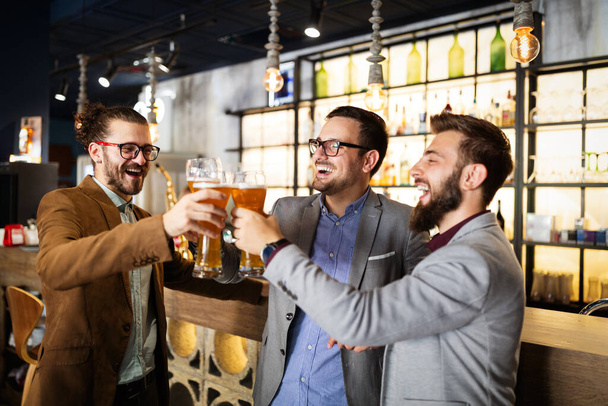 People, men, leisure, friendship and celebration concept. Happy business male friends drinking beer and clinking glasses at bar or pub - Fotoğraf, Görsel