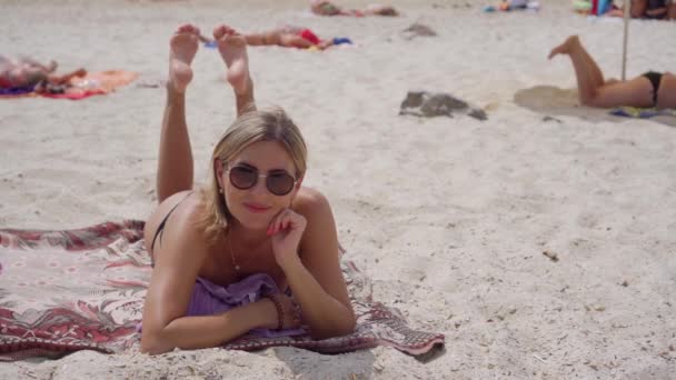 Een vrouw met een bril van de zon ligt op het zandstrand. Ze rust zorgeloos en zwaait met haar benen. - Video