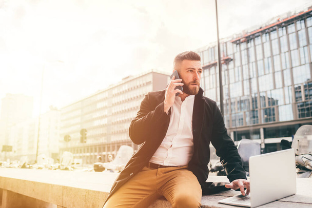 Young businessman outdoor talking smartphone using computer - Caucasian bearded man remote working having business call - business, technology, working concept - Foto, imagen