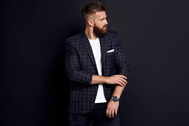 Handsome man with perfect hairdo and beard adjusting sleavs while standing against black background. - Foto, Bild