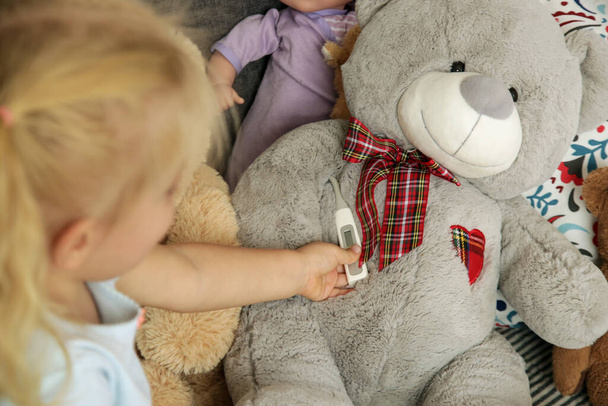 Adorable blonde toddler girl playing with a thermometer and looking after her sick teddy bear.  - Photo, image