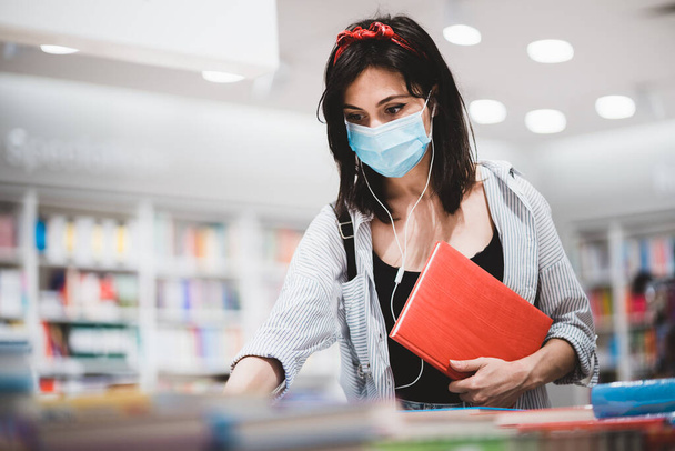 Moda kıyafetleri içinde genç bir kız öğrenci sınava hazırlanıyor ve kütüphanede kitap arıyor. Coronavirus salgını sırasında üniversitede okuyan yüzü tıbbi maskeli sevimli hippi kadın. - Fotoğraf, Görsel
