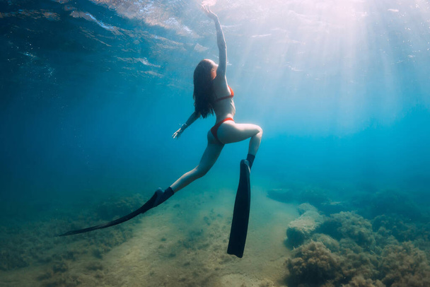 Λεπτή γυναίκα ελεύθερη δύτης ποζάρουν κάτω από το νερό σε μπλε θάλασσα με ακτίνες του ήλιου. Ελεύθερη κατάδυση με πτερύγια στη θάλασσα - Φωτογραφία, εικόνα