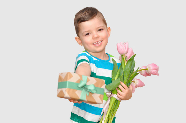 A cute little boy gentleman in a colored striped t-shirt holding a bouquet of pink tulips and a gift box with a green bow. - Photo, Image