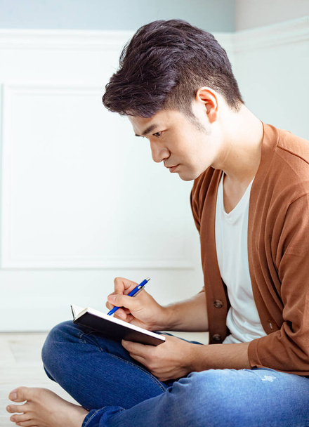 Cross-legged young asian man writing in notebook. - Valokuva, kuva