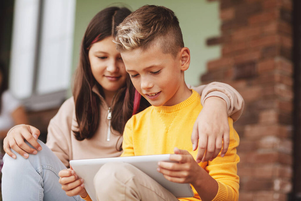 Bruder und Schwester sitzen nach der Schule auf den Stufen und spielen auf einem digitalen Tablet. Moderne Kinder, - Foto, Bild