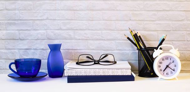 Against the background of a light brick wall, on a light desktop, there are diaries, a white alarm clock, glasses, a blue cup, a blue vase and pencils. Close-up of the workplace. - 写真・画像
