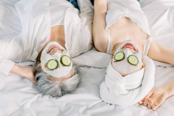 Close up top image of women, senior gray haired lady mother and her young pretty daughter having fun in the bedroom and making clay facial masks with cucumber covering their eyes. Beauty skin care - Φωτογραφία, εικόνα