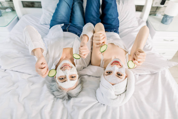 Top view portrait showing two women, senior pretty grandmother and young smiling granddaughter, lying on the bed at home, with facial masks on face, holding cucumber slices. Family spa - Photo, image