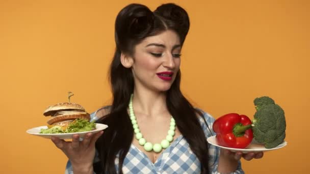 Χαμογελώντας pin up γυναίκα κοιτάζοντας τα πιάτα και δεν μπορεί να επιλέξει μεταξύ fast food και λαχανικά - Πλάνα, βίντεο