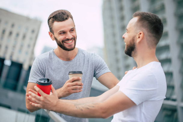 Two handsome young bearded best friends in casual clothes are standing outdoor with coffee cups laughing and have a conversation. Coffee break, startup, coworking, business. - Foto, Imagen
