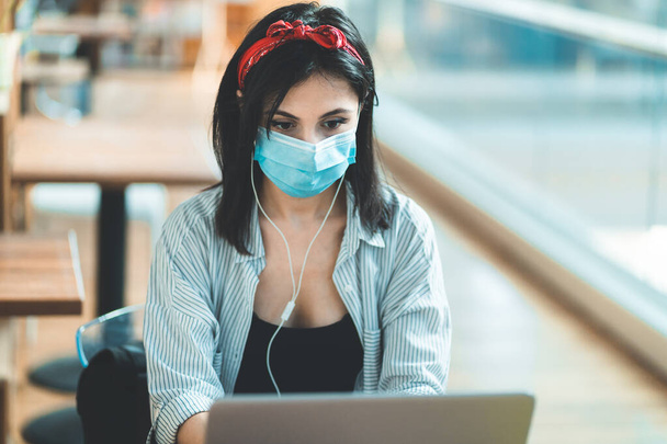 Молода жінка носить захисну маску для обличчя, використовує ноутбук і навчається в університетській бібліотеці. Жінка працює віддалено від кав'ярні або бібліотеки
 - Фото, зображення