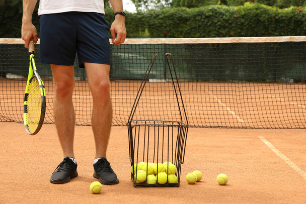 Man hold racket on clay court with basket of tennis balls - Фото, изображение