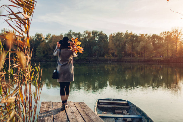 Stylish woman walking on lake pier by boat admiring autumn landscape in forest holding leaves. Fall season activities - Photo, Image