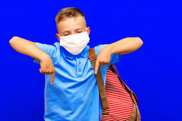 Молодий школяр носить маску в синій сорочці рюкзак, спрямований пальцями рук на робочий простір ізольований на яскраво-синьому фоні
 - Фото, зображення