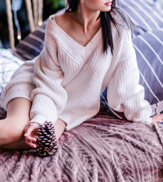 Chica morena se sienta en una cama suave en un suéter blanco con un cono de pino en las manos. Foto vertical - Foto, Imagen