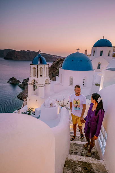 Santorini Kreikka, nuori pari luksuslomalla Santorinin saarella katselemassa auringonnousua sinisen kupolin kirkon ja Oia Santorinin kalkitun kylän luona Kreikka auringonnousun aikana - Valokuva, kuva
