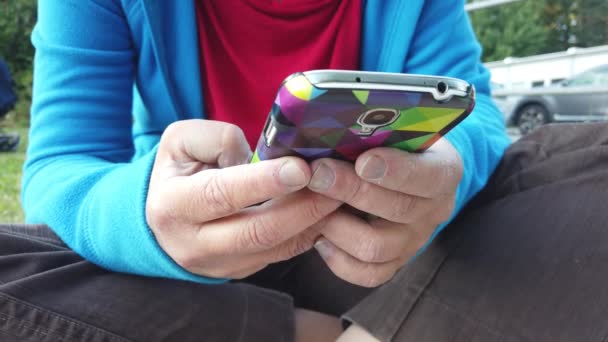 μια νεαρή γυναίκα χέρια καθώς κοιτάζει το κινητό της τηλέφωνο κάθεται σε ένα γκαζόν - Πλάνα, βίντεο